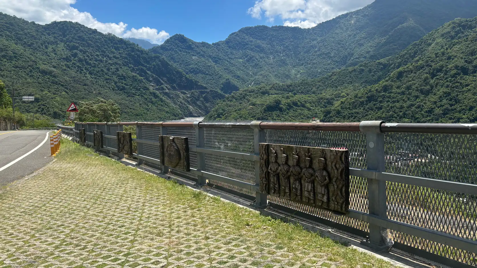 山區公路旁的橋樑欄杆上鑲著原住民人像木雕浮雕，背後是南臺灣層疊的青翠山巒與溪谷
